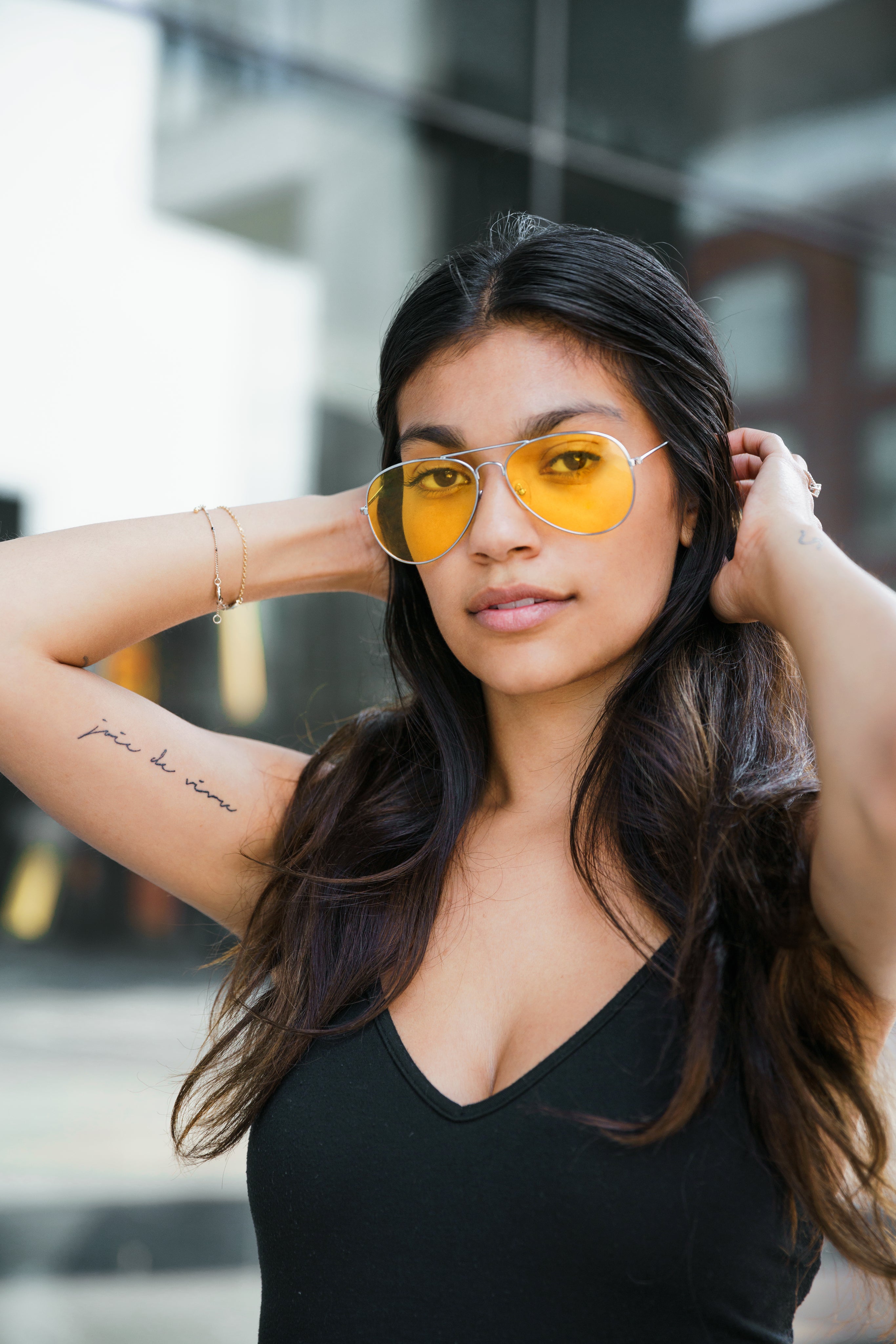 Confident woman wearing yellow aviator sunglasses and a black top, standing outdoors with urban background